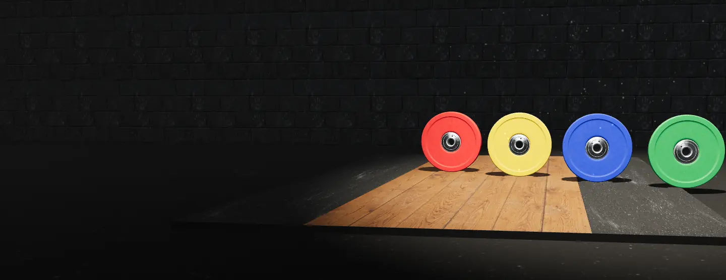 Red, yellow, blue and green bumper plates standing in a row on a gym floor under a spotlight